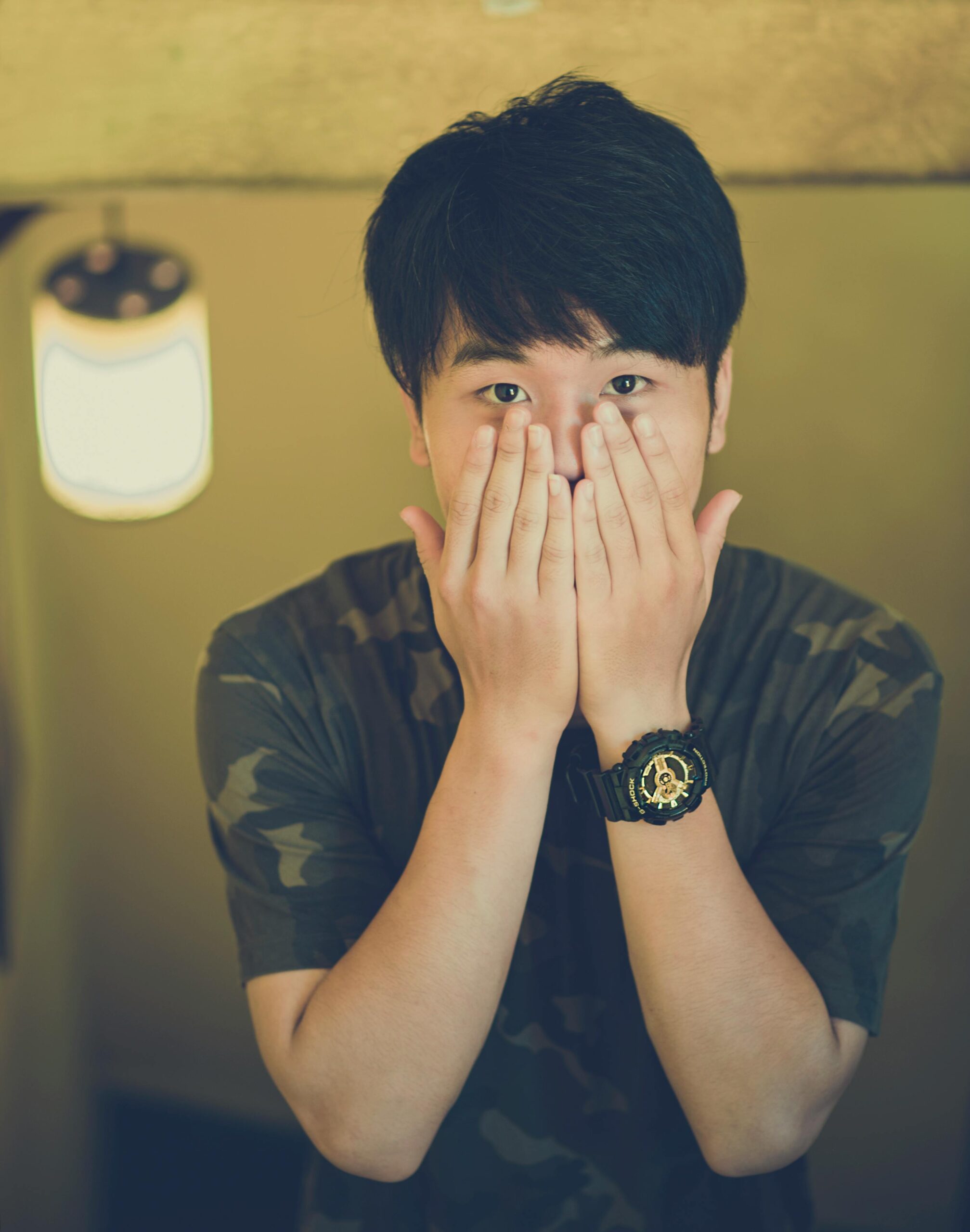 A surprised young man covers his face with his hands wearing a camouflage shirt indoors.